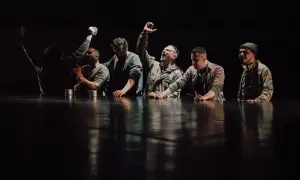 Grupa mężczyzn w ciemnych ubraniach, z rękami na stole, na tle czarnej sceny.