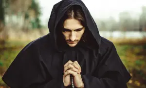 Mężczyzna w czarnym kapturze modli się, trzymając w dłoniach paciorki różańca.