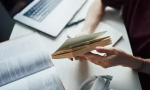 Osoba trzymająca otwartą książkę, obok laptop i słuchawki.