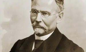 Portret mężczyzny w okularach i z wąsami, ubranego w garnitur.