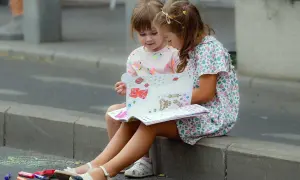 Dwie dziewczynki siedzą na krawężniku i oglądają książkę z naklejkami.