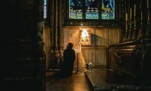 Osoba klęczy przed ikoną w ciemnym kościele, zapalając świece.