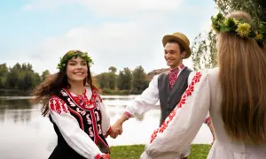 Trzy osoby w tradycyjnych strojach ludowych trzymają się za ręce, tańcząc na tle jeziora.