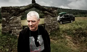 Mężczyzna z siwymi włosami przed kamiennym łukiem, za nim czarny SUV z bagażnikiem dachowym.