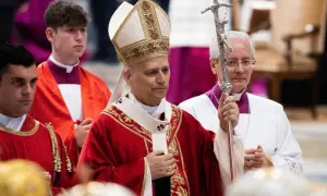 Biskup w stroju pontyfikalnym trzyma pastorał, obok klerycy w czerwonych szatach.