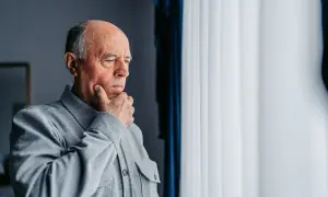 Starszy mężczyzna w szarej koszuli zamyślony przy oknie.