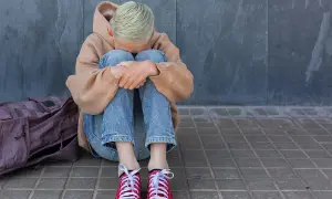 Młoda osoba w beżowej bluzie z kapturem i jeansach siedzi skulona na chodniku obok plecaka.