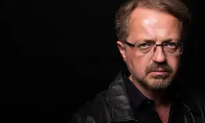 Mężczyzna w okularach i skórzanej kurtce patrzy poważnie na wprost na czarnym tle.