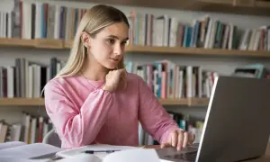 Dziewczyna w różowej bluzie uczy się przy laptopie na tle regałów z książkami.