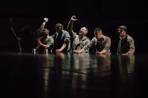 Grupa mężczyzn w ciemnych ubraniach, z rękami na stole, na tle czarnej sceny.