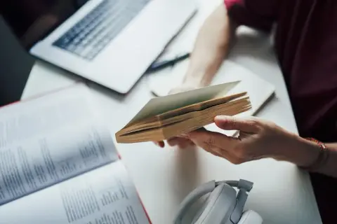 Osoba trzymająca otwartą książkę, obok laptop i słuchawki.