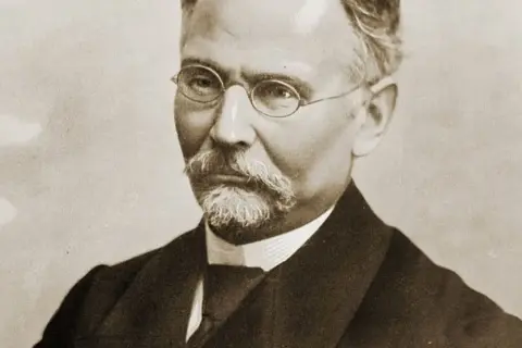 Portret mężczyzny w okularach i z wąsami, ubranego w garnitur.