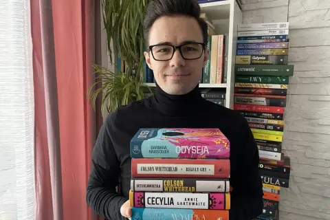 Mężczyzna w czarnym golfie trzyma stos kolorowych książek.