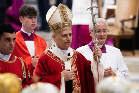 Biskup w stroju pontyfikalnym trzyma pastorał, obok klerycy w czerwonych szatach.