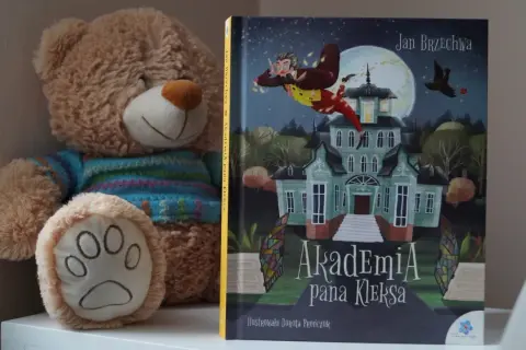 Pluszowy miś obok książki "Akademia Pana Kleksa" z ilustracją latającego Ambrożego Kleksa.