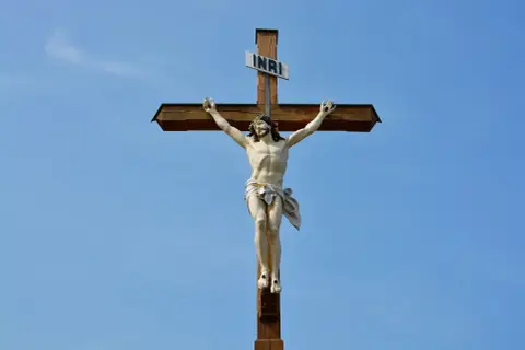 Figurka Jezusa na drewnianym krzyżu z tabliczką INRI na tle błękitnego nieba.