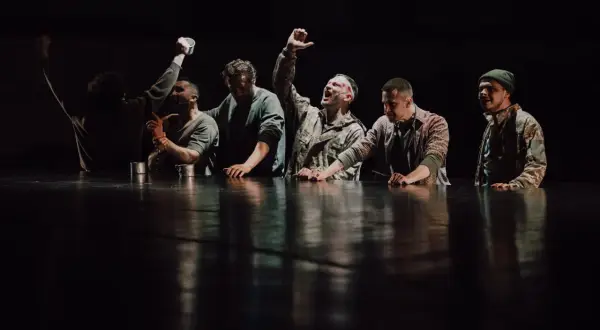 Grupa mężczyzn w ciemnych ubraniach, z rękami na stole, na tle czarnej sceny.