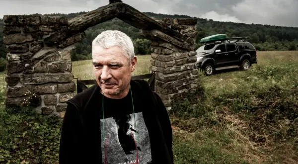 Mężczyzna z siwymi włosami przed kamiennym łukiem, za nim czarny SUV z bagażnikiem dachowym.