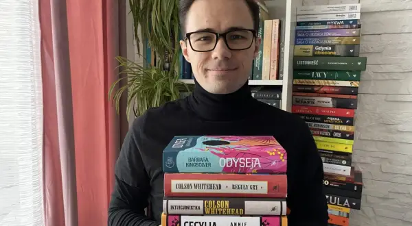 Mężczyzna w czarnym golfie trzyma stos kolorowych książek.