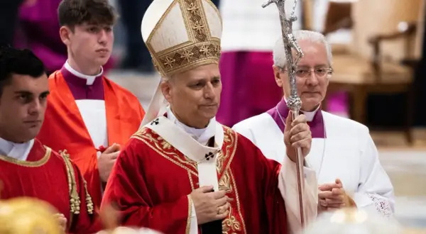 Biskup w stroju pontyfikalnym trzyma pastorał, obok klerycy w czerwonych szatach.