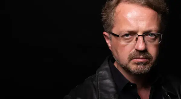 Mężczyzna w okularach i skórzanej kurtce patrzy poważnie na wprost na czarnym tle.