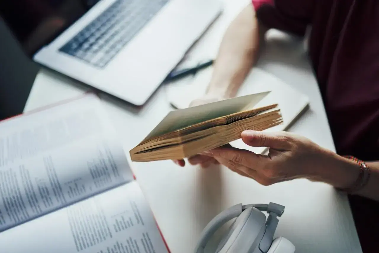 Osoba trzymająca otwartą książkę, obok laptop i słuchawki.