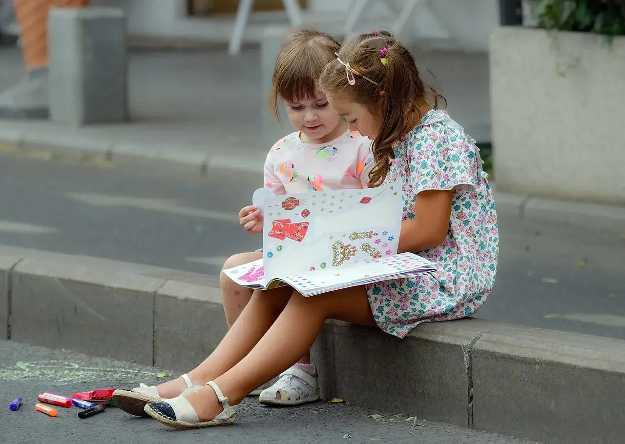 Dwie dziewczynki siedzą na krawężniku i oglądają książkę z naklejkami.