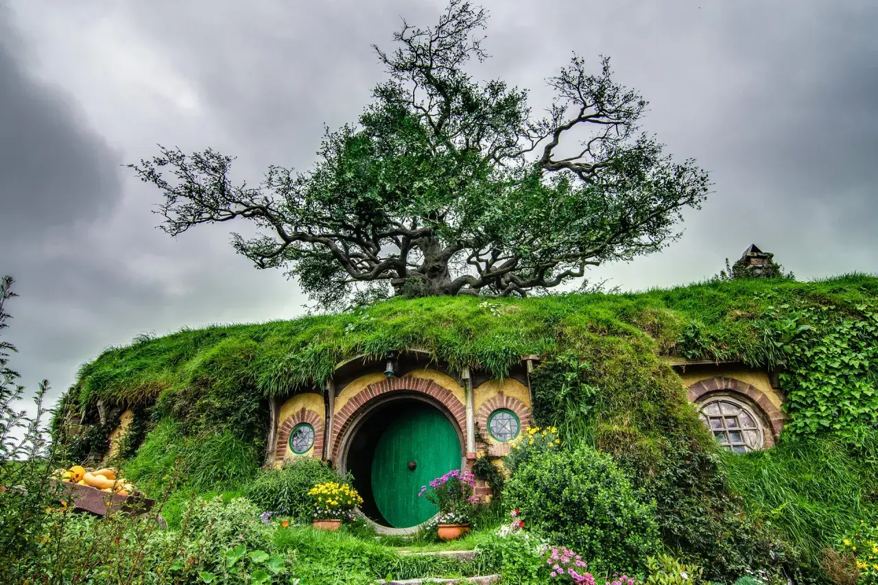 Zielone drzwi hobbitowej nory pod drzewem, otoczone trawą i kwiatami.