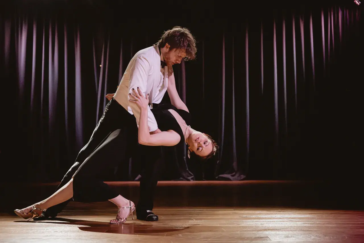 Para tańczy tango na scenie, kobieta w czarnej sukni w głębokim ukłonie, mężczyzna w białej marynarce.