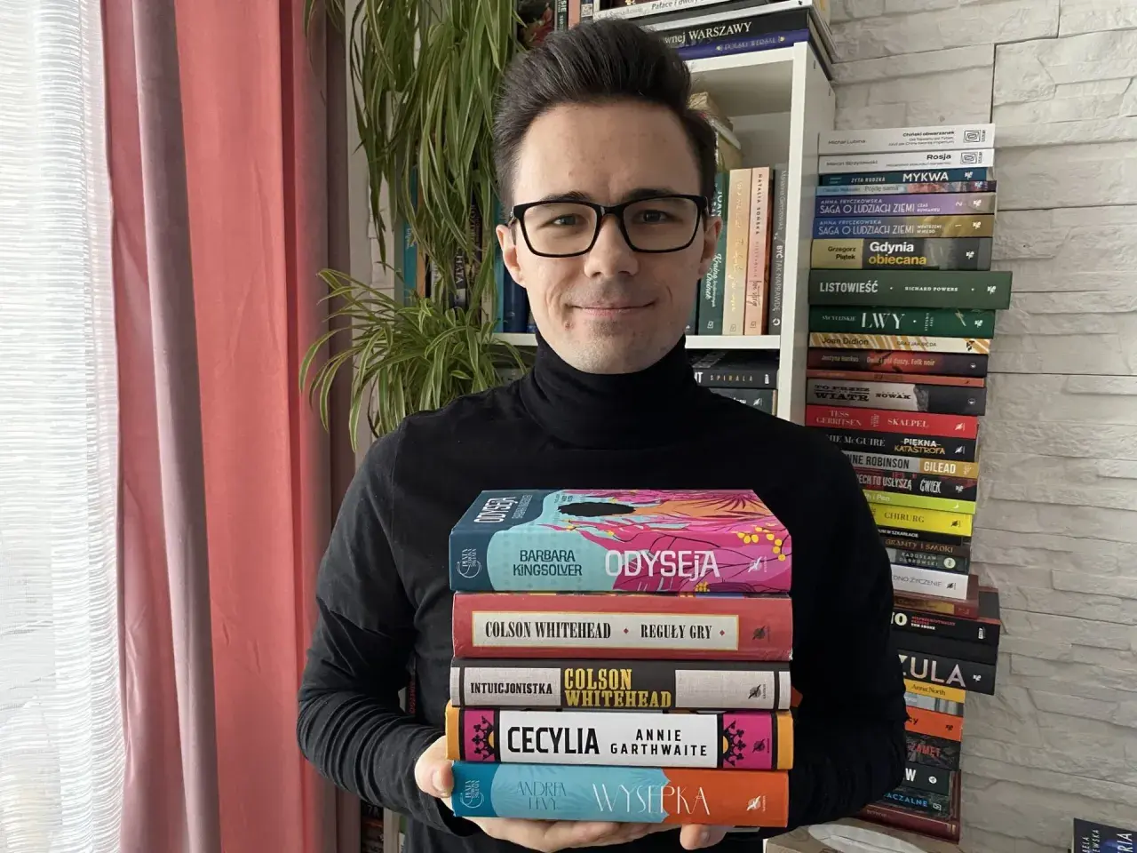Mężczyzna w czarnym golfie trzyma stos kolorowych książek.