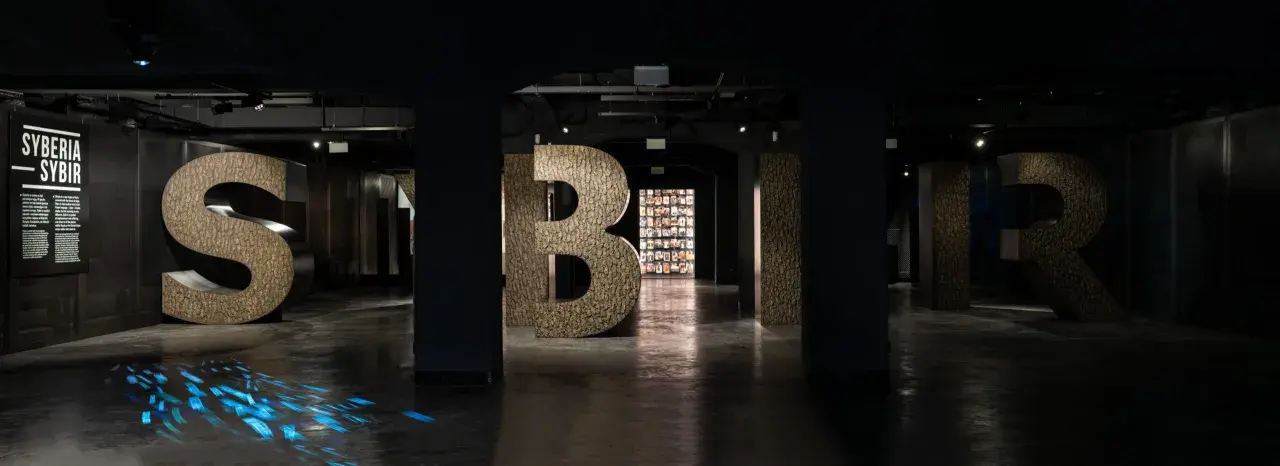 Wielkie litery "S", "Y", "B", "I", "R" w ciemnym wnętrzu, z niebieskimi refleksami na podłodze.