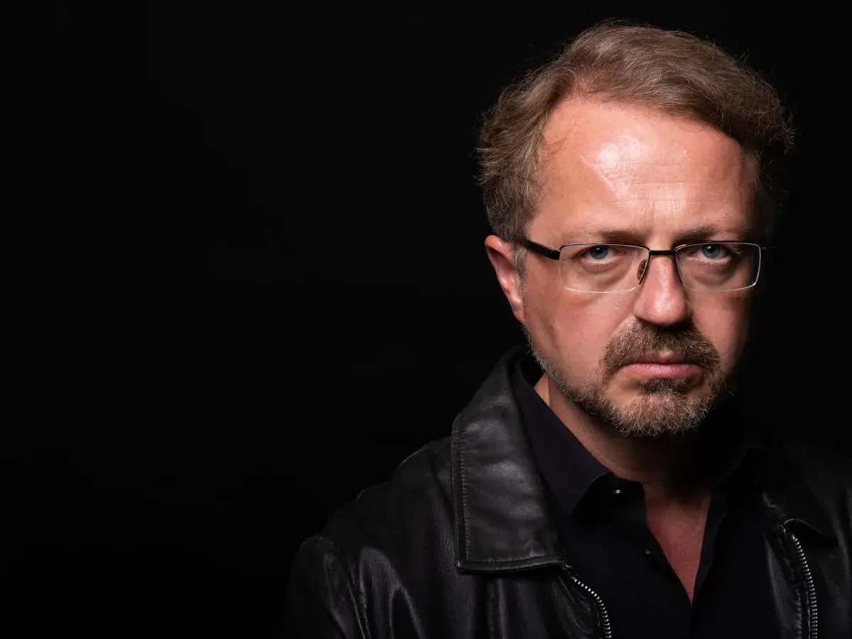 Mężczyzna w okularach i skórzanej kurtce patrzy poważnie na wprost na czarnym tle.
