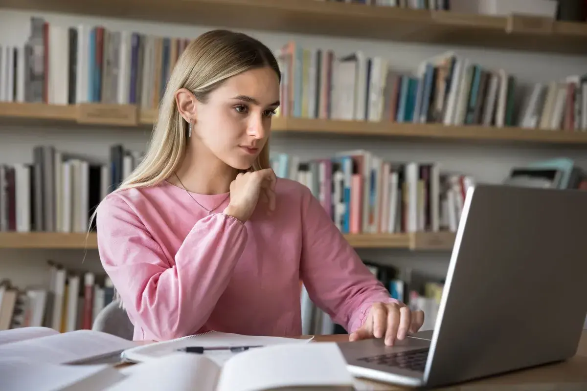 Dziewczyna w różowej bluzie uczy się przy laptopie na tle regałów z książkami.