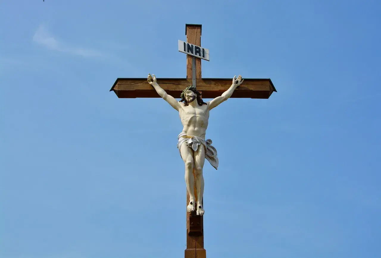 Figurka Jezusa na drewnianym krzyżu z tabliczką INRI na tle błękitnego nieba.