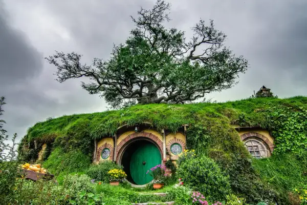 Zielone drzwi hobbitowej nory pod drzewem, otoczone trawą i kwiatami.