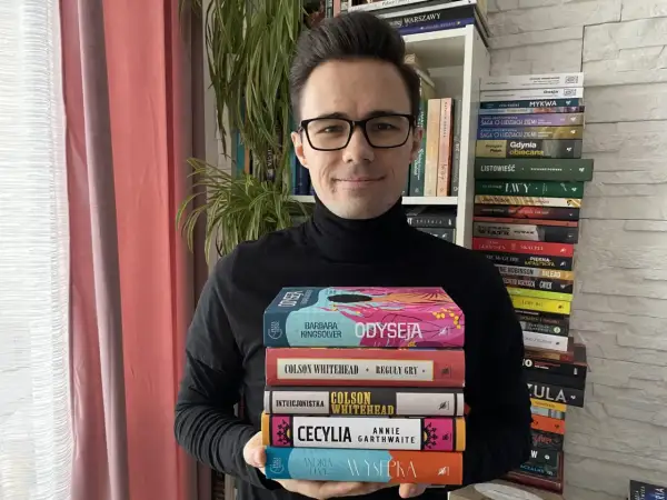 Mężczyzna w czarnym golfie trzyma stos kolorowych książek.