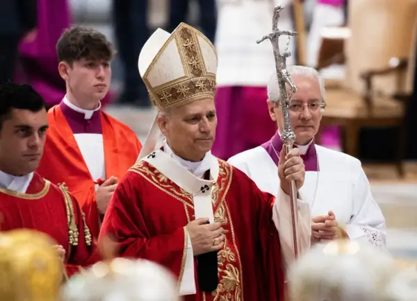 Biskup w stroju pontyfikalnym trzyma pastorał, obok klerycy w czerwonych szatach.