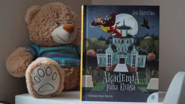 Pluszowy miś obok książki "Akademia Pana Kleksa" z ilustracją latającego Ambrożego Kleksa.